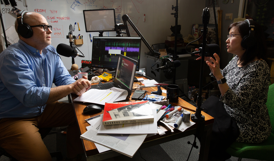 A man sits in front of two screens at a cluttered desk, listening to a woman speaking into a microphone on the other side of the desk.