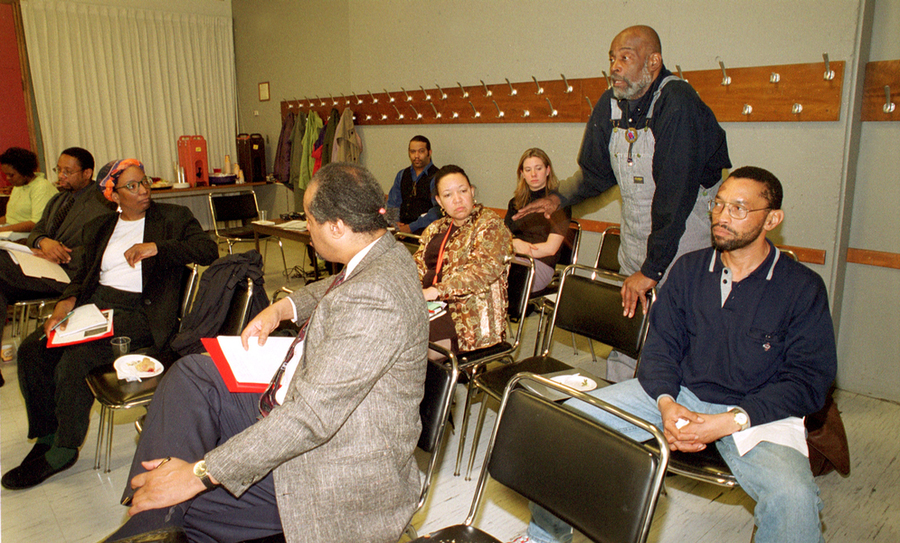 About nine people sit in a classroom. Mel King is standing near the back speaking, and people are turned to listen to him.