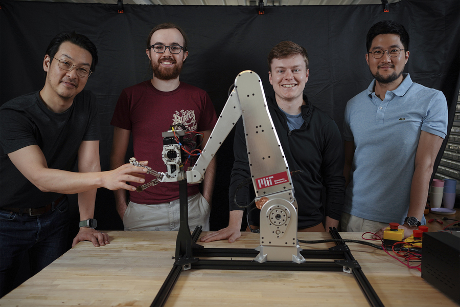 Sangbae Kim, Elijah Stanger-Jones, Andrew SaLoutos, and Hongmin Kim pose for a portrait as they stand behind a metal robot gripper that resembles an excavator. Sangbae Kim’s fingers lace with the gripper.