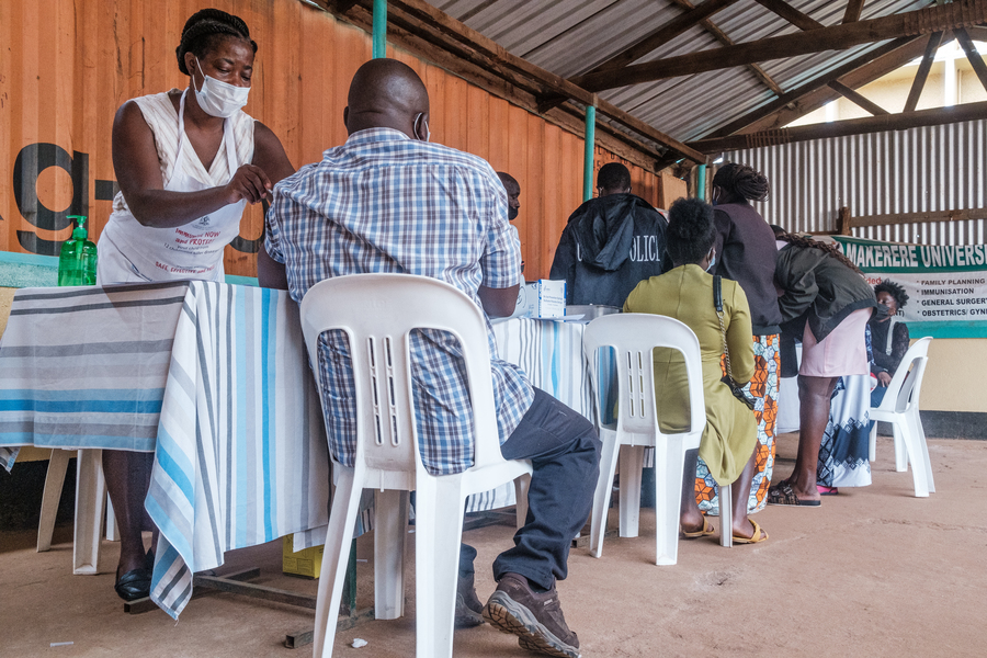 Several people, seen from the back, sit in plastic chairs at a table while health care workers inject vaccine.
