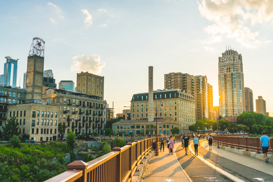 People walk on a pedestrian walkway in front of a city skyline at sunset.