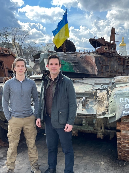 Ian Miller and Evan Platt stand in front of a battered tank. A Ukranian flag flies from the tank.