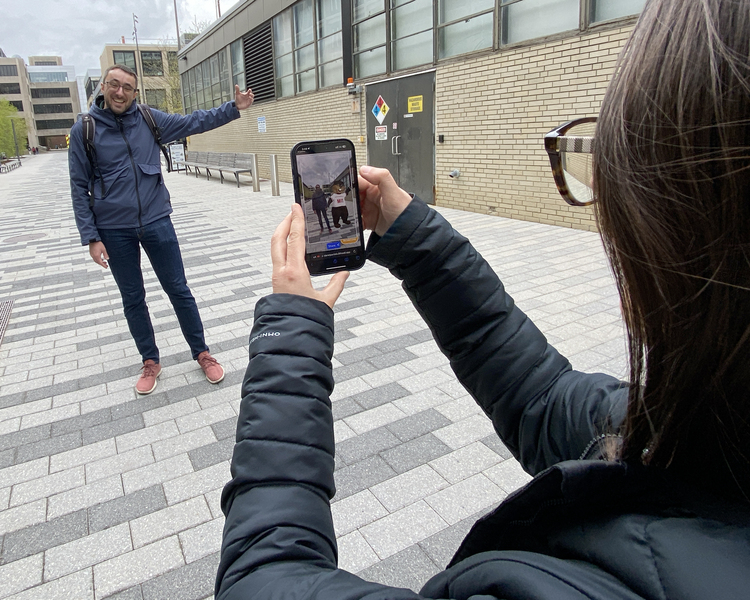 A person holds a phone toward another person posing with their arm in the air, as if around an invisible person’s shoulder