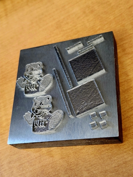Several silver, metallic shapes sit on top of a square of the same material. The shapes include a pair of MIT beaver mascots, plus a variety of squares, rods, and cubes.