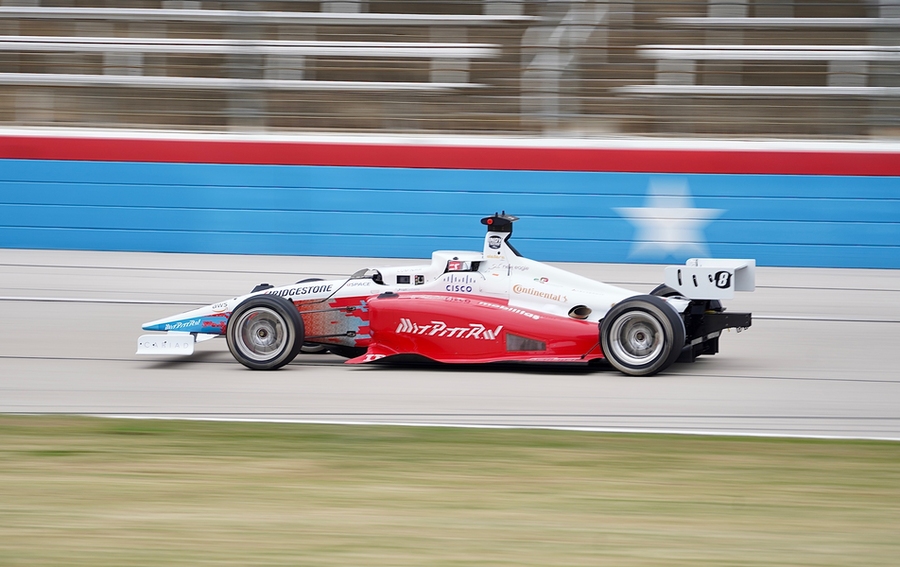 A red-and-white driverless race car appears to stand still as the background blurs past it. MIT-PITT-RW is written on the side, accompanied by sponsor logos.