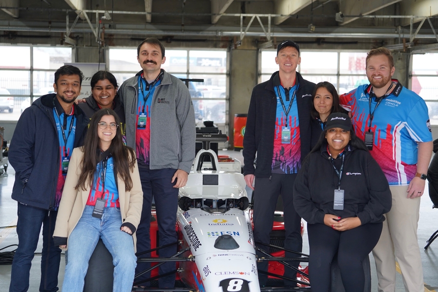 Eight people pose with their autonomous race car, photographed from the front, with four on each side. 