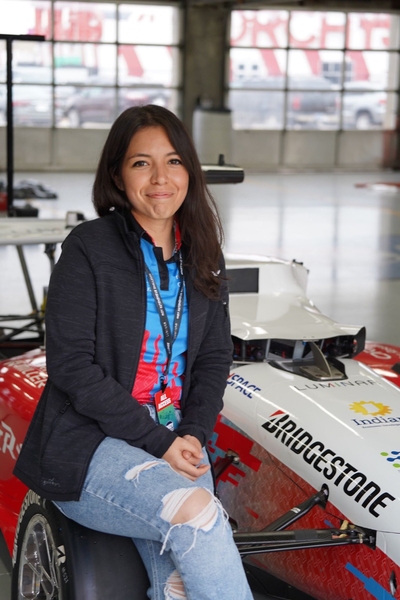 Cindy Heredia poses and smiles at the camera, sitting on her teams’ white, blue, and red driverless race car at the Indy Autonomous Challenge