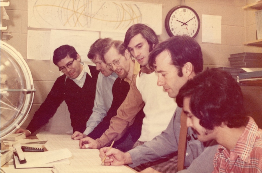 Six men at SAS control room desk. Five are watching the sixth write with a pencil on paper.
