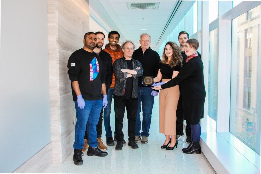 Eight people pose standing in a white hallway with the HUMANS nanowafer held between them. On their left is a wall, and the right are windows.