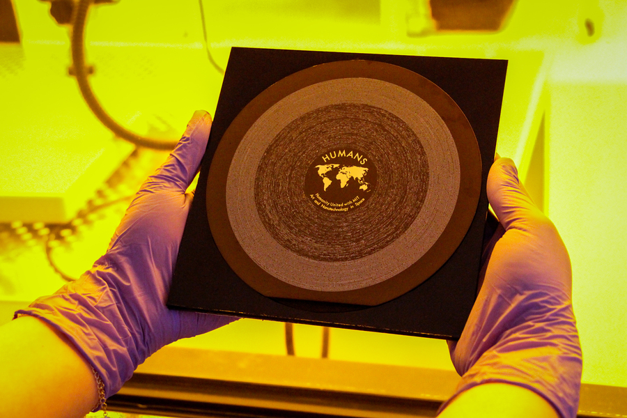 Two hands in blue gloves hold the HUMANS nanowafer. The wafer is 6 inches in diameter, is a brown/gold color, and has an equal-area projection map etched into the center, along with “HUMANS” and “Humanity United with MIT Art and Nanotechnology in Space.” Around the map and words are groves, like a record. Yellow light from the MIT.nano lab is in the background.