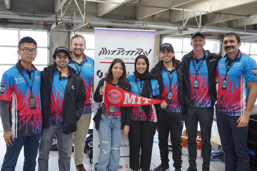 Eight people in identical team polo shirts and lanyards stand in a line with their banner in the background. Two are holding an MIT pennant.