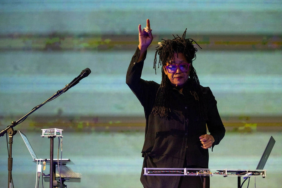 Pamela Z performing. She is gesturing to electronic instruments in front of her, with a pale green and brown striped backdrop behind her.