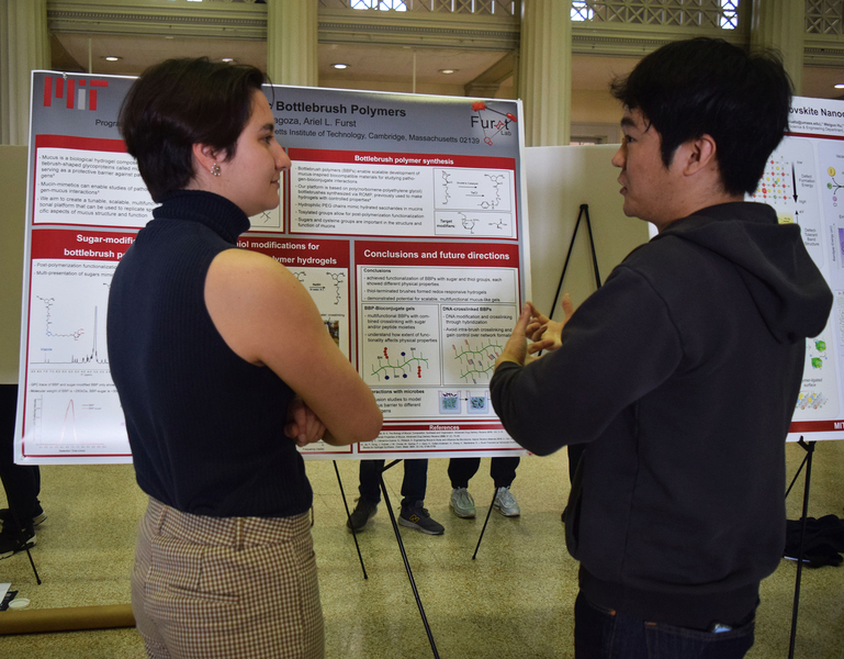 Two people seen in profile converse in front of a poster