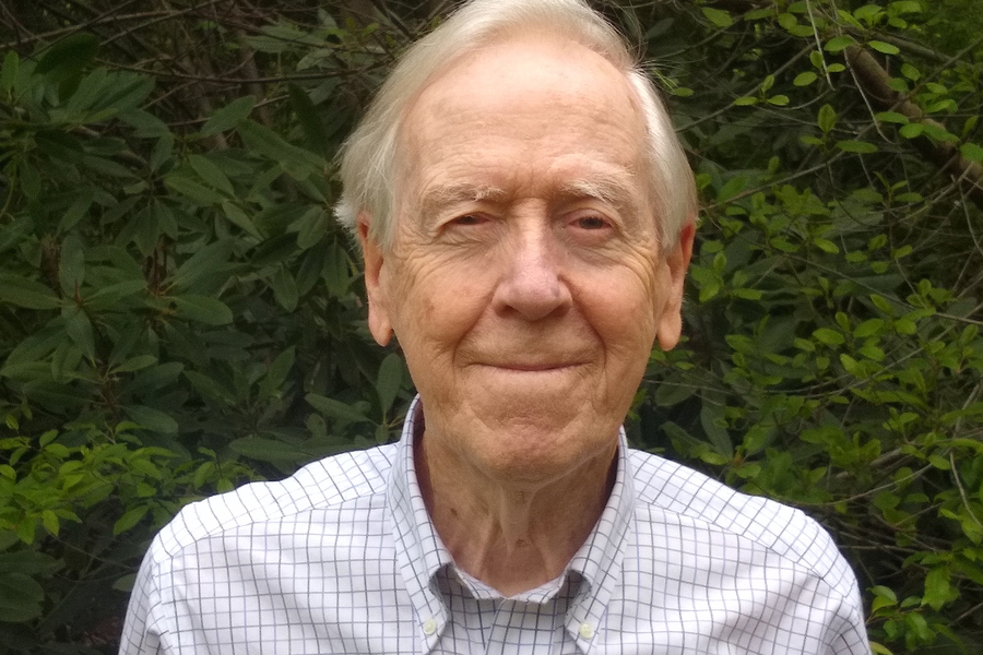 Headshot of a white-haired Gil Strang, who stands before a lush collection of green shrubs.