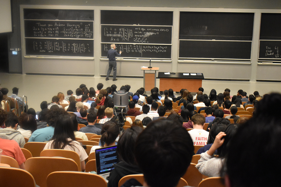 Before a large crowd of students seated in rows, a white-haired Gil Strang writes at a blackboard.