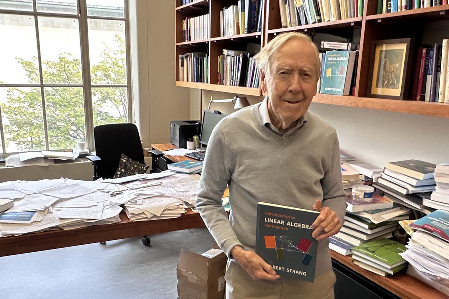 A white-haired Gil Strang holds up a copy of his textbook, “Introduction to Linear Algebra,” in an office filled with books.