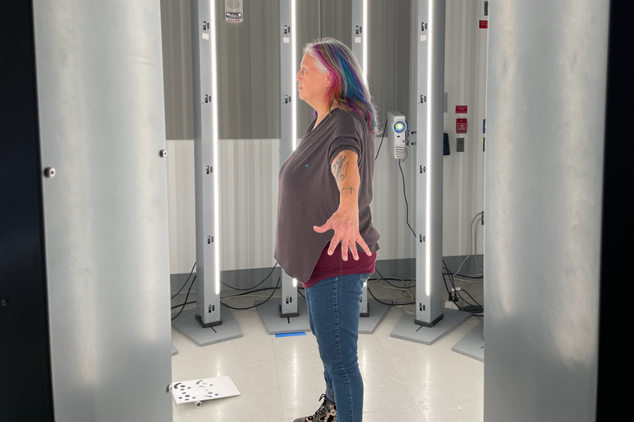 A person with long, multicolored hair standing with their arms spread inside a 3D scanner