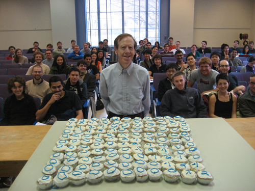 Gil Strang in middle age stands before an 11 by 11 array of vanilla cupcakes with white frosting that also have numbers on them written in blue. Behind him are several rows of students seated in classroom chairs, smiling for the camera. 