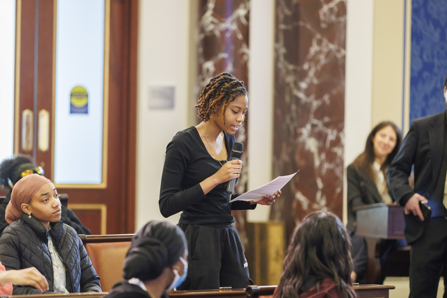 Photo of a middle school girl standing behind a desk in a replica of the U.S. Senate Chambers, speaking into a microphone and reading from a piece of paper. Several students and adults look on.