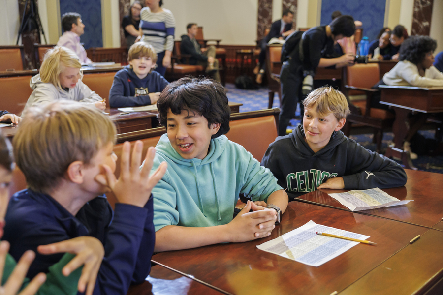 Photo of 3 middle school boys in an animated discussion about the paper assignement sitting on the desk in front of them. A dozen students sit in the rows of desks behind them.
