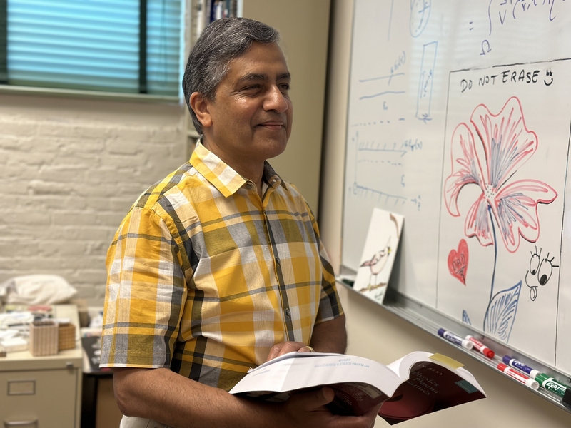 Abhay Ram stands in front of a whiteboard, holding an open physics textbook.