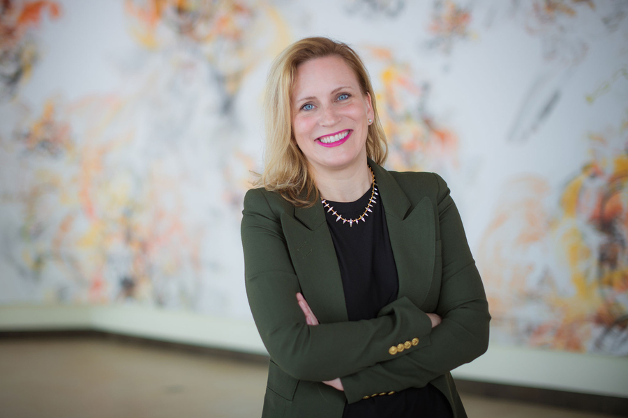 Angela Koehler stands with arms folded in front of white wall with colorful design.