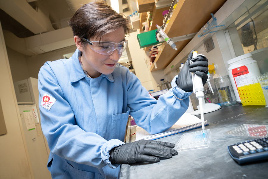 Farquhar wears a blue lab coat, black gloves, and safety goggles inside the lab. Their hand rests on a counter with lots of supplies, including vials and a calculator.
