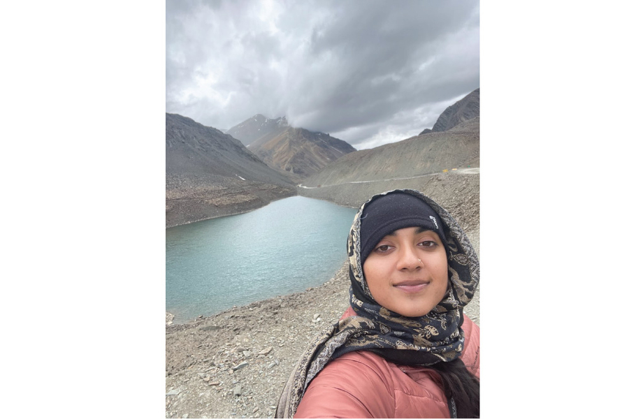Selfie of Neosha Narayanan wearing a beanie with a loosely wrapped patterned hijab, with a glacial lake and cloud-shrouded mountaintop in the background