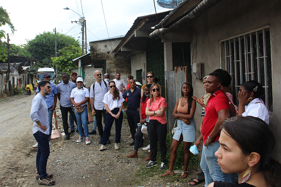 About 2 dozen people are gathered togehter in an underdeveloped community in Colombia. They stand beside simple cinderblock dwellings with corrugated tin roofs that line a dirt road.