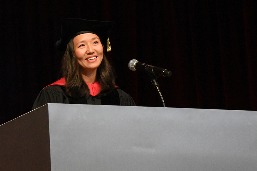 Michelle Wu, wearing graduate cap and gown, speaks at a podium before a black background