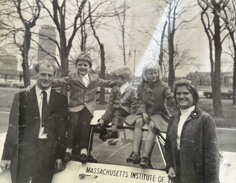 In a battered sepia toned photo from approximately the 1960s, a young family, dressed in sharp formalwear, pose around an electric car.
