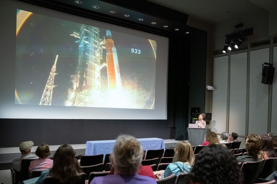 People in a theater watch a video of a rocket launch projected on a screen. 