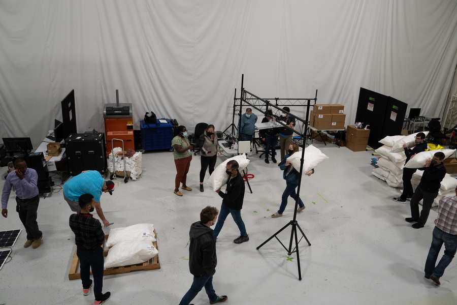 Inside an indoor warehouse set up with rigs and stacks of large rice bags, people carry barcoded and RFID tagged bags past a scanner system and hold their cellphones close to scan them.