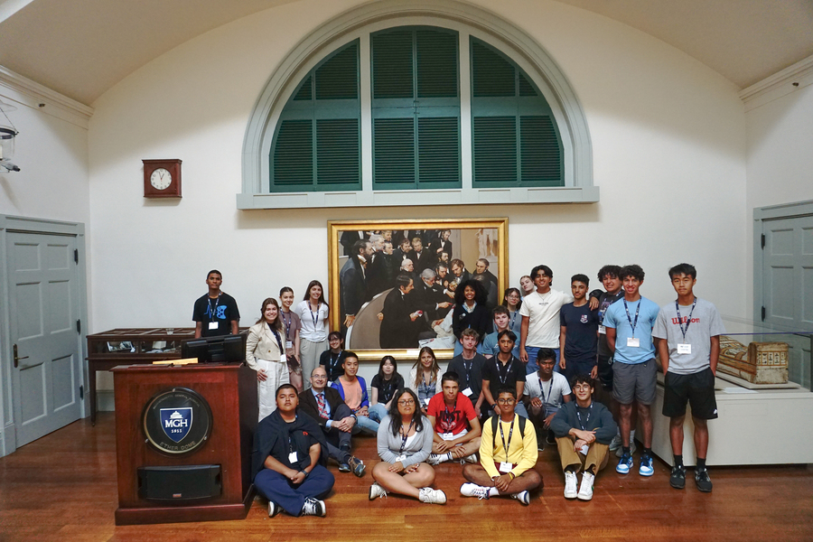 26 people, some standing, some sitting, pose in front of a painting in an auditorium. 