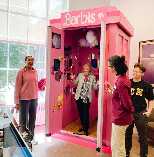 Sally Kornbluth is standing in a pink Barbie-themed tardis labeled "Barbis," wearing pink 80s sunglasses and holding up jazz hands. One student stands at left and two students stand at right. Everyone in the scene is laughing.