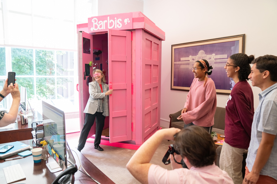 alt text: Sally Kornbluth at the entrance to a Barbie-themed tardis, labeled Barbis. She wears pink 80s-style sunglasses. There are three students on the right. Everyone in the scene is laughing. Someone in the foreground crouches to take a photo and a hand holding a cellphone to take a pic is at left.