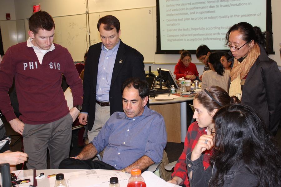 Five people around a table listen to Edward Crawley, seated. Four others surround a table in the background. 