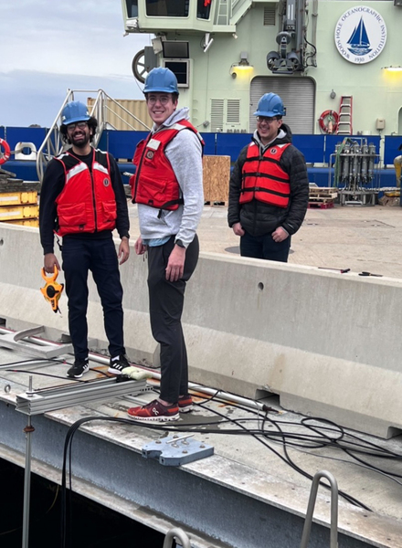 Three team members wear life jackets and hard hats while outside in a work area.