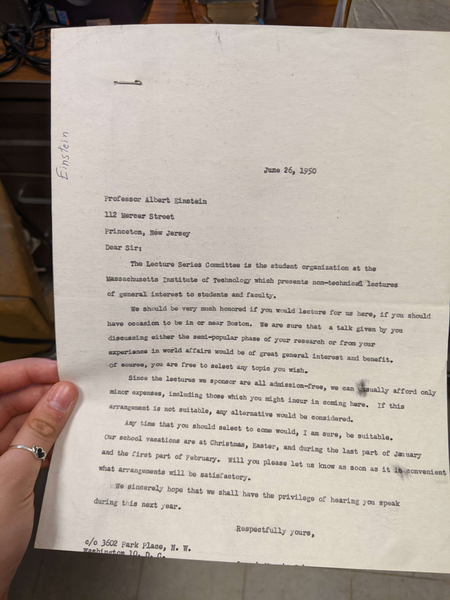 A hand holds up a typed letter to Albert Einstein dated June 26, 1950, from the Lecture Series Committee at MIT, inviting him to speak at MIT. The sender's name is not visible.