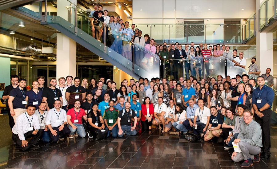 Hundreds of MIT Bootcamps alumni pose in a large atrium, near stairs. 