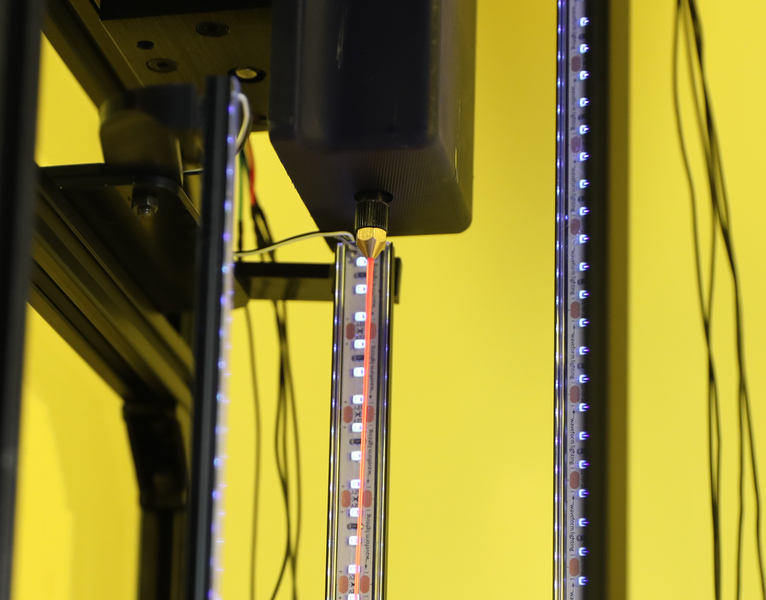 Coming out of the machine’s nozzle is an orange resin fiber. The black machine has strips of LEDs.