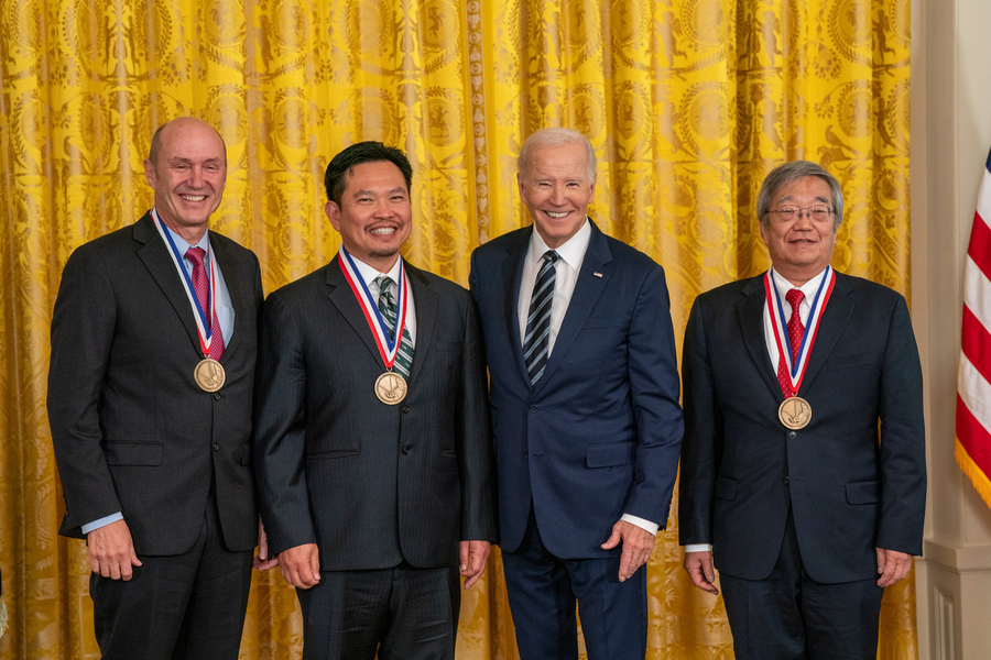 From left to right: Eric Swanson, David Huang, President Joe Biden, and James Fujimoto stand against gold curtains for a photo. The winner wear large gold medals around their necks.