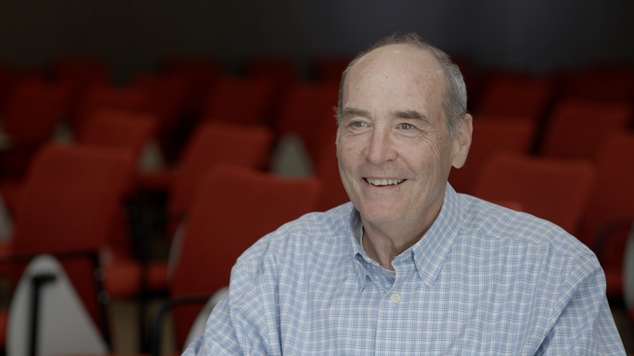 Mark Bear is sitting in an auditorium full of red chairs and smiling.