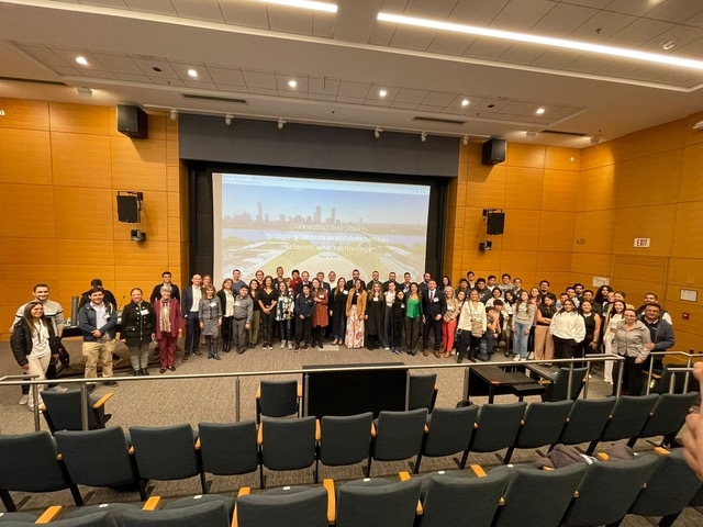 About 65 people pose standing in a semicircle on a large auditorium stage, with the backs of empty seats in the foreground and a video screen behind them.