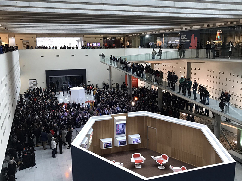 A large crowd photographed from a catwalk is gathered for the exhibit's opening reception. An overhead of the Cybersyn Operations Room is in the foreground.
