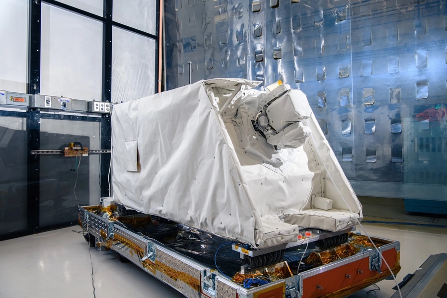 A refrigerator-sized structure with a white covering sits on a platform in a cleanroom