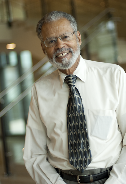 Portrait photo of Willard Johnson, who stands indoors with shiny items on a wall behind him out of focus