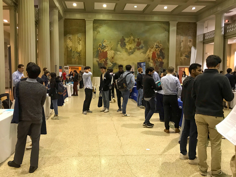 A few dozen people mill about between two rows of tables at a career fair on the MIT campus.
