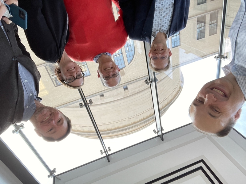 Five people stand in front of a window at MIT.nano; in the background rises the MIT dome. The photographer is presumably kneeling as the subjects are all looking down at the camera.