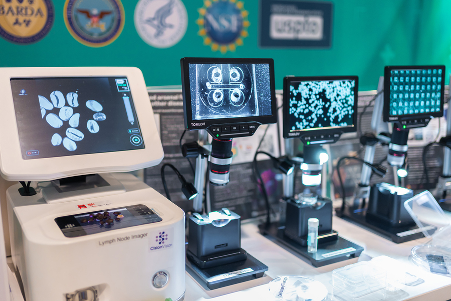 Four microscopes equipped with digital screens are seen close up on a table. Nanoscale cells are seen in greyscale on each of the screens